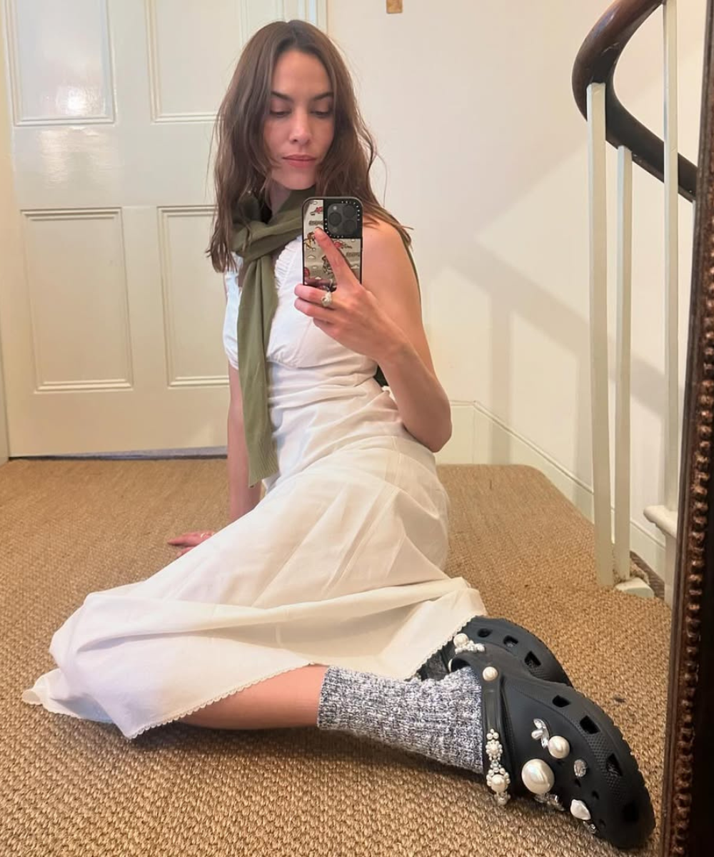 Pessoa sentada no chão, vestindo vestido branco e Crocs decorados. Segura o celular para tirar foto.