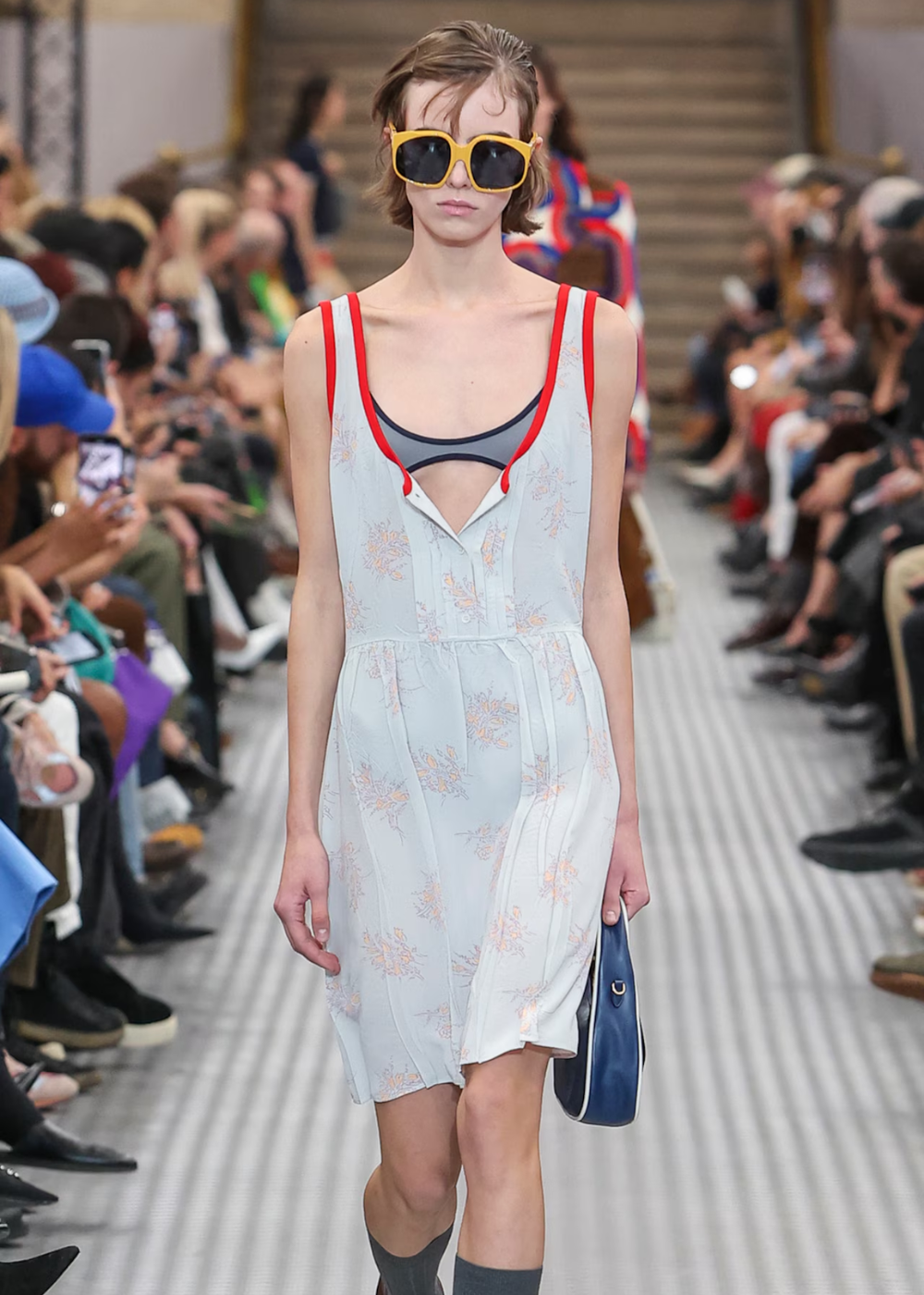 Modelo desfila com vestido claro, detalhes florais e óculos grandes. Sugestiva de um vestido favorito na passarela de moda.