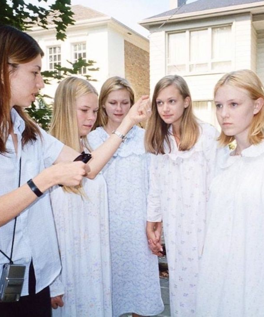Quatro jovens de mãos dadas em vestidos leves, ao ar livre, recebendo instruções de uma mulher.