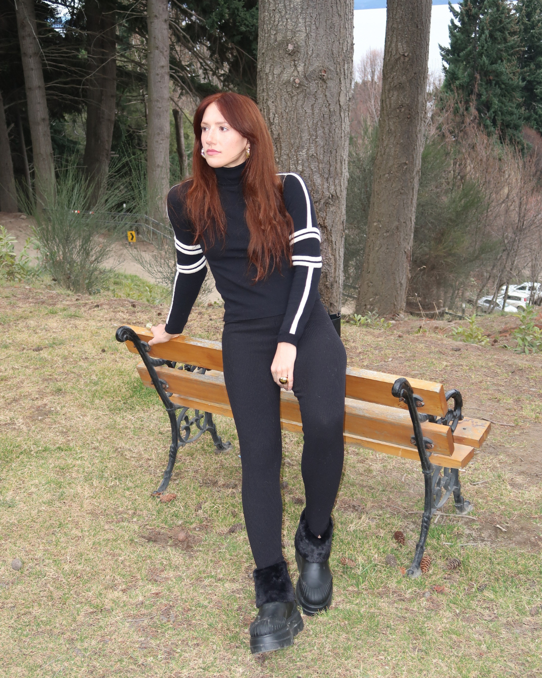 Mulher de cabelo castanho usa roupa preta e botas com pele, apoiada em um banco no parque. Tendências de alto inverno