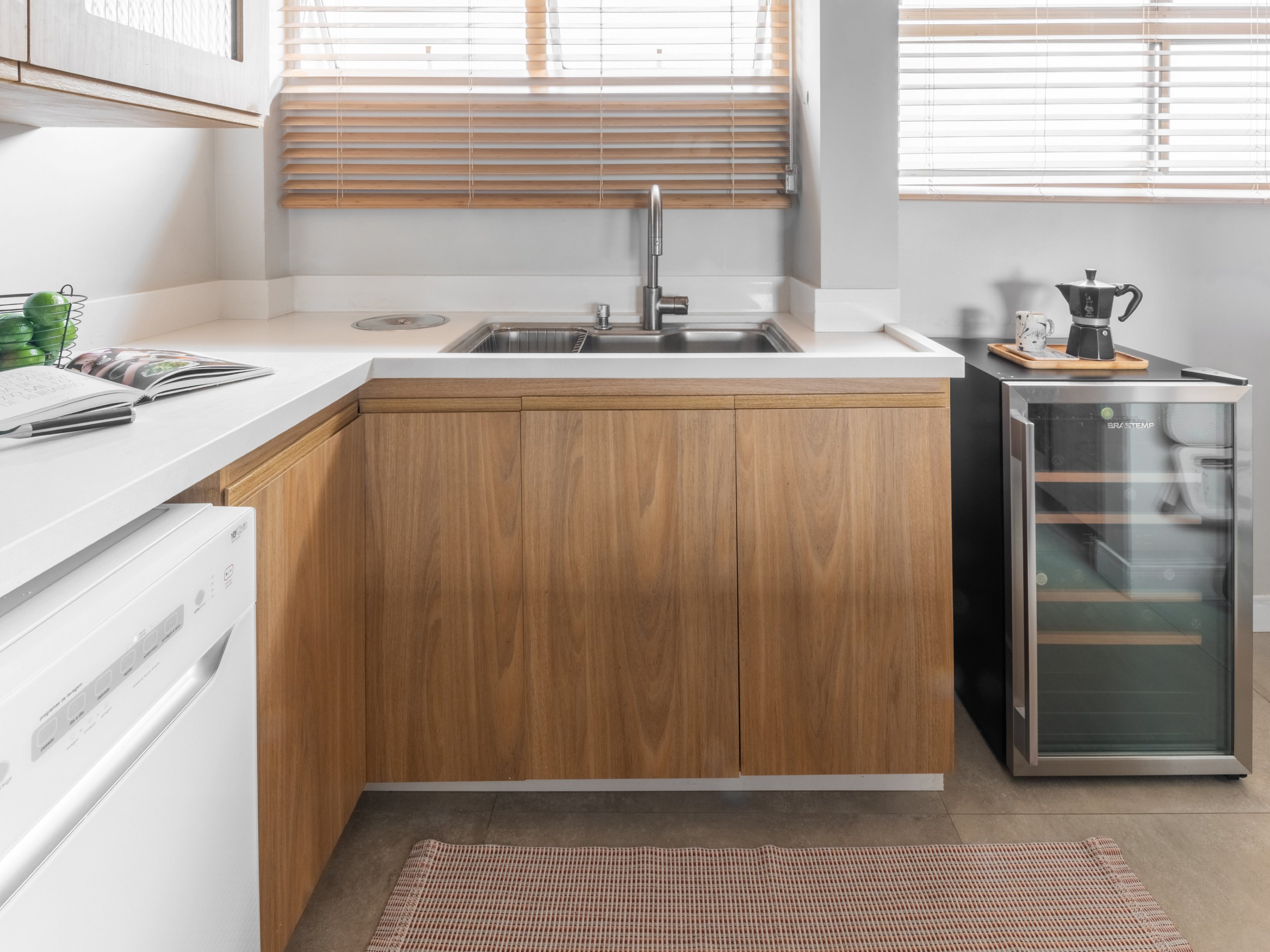 Cozinha com cuba de aço inox, armários de madeira clara e adega. Cortinas de madeira nas janelas.