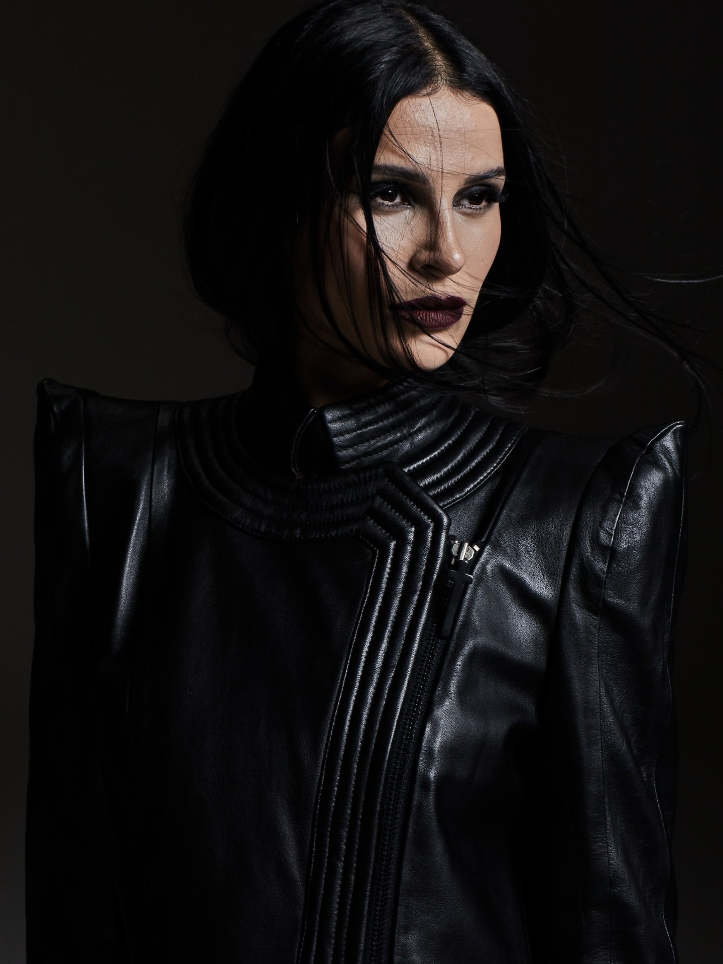Mulher com cabelo escuro e jaqueta de couro preto, olhando para o lado, em pose confiante e forte.