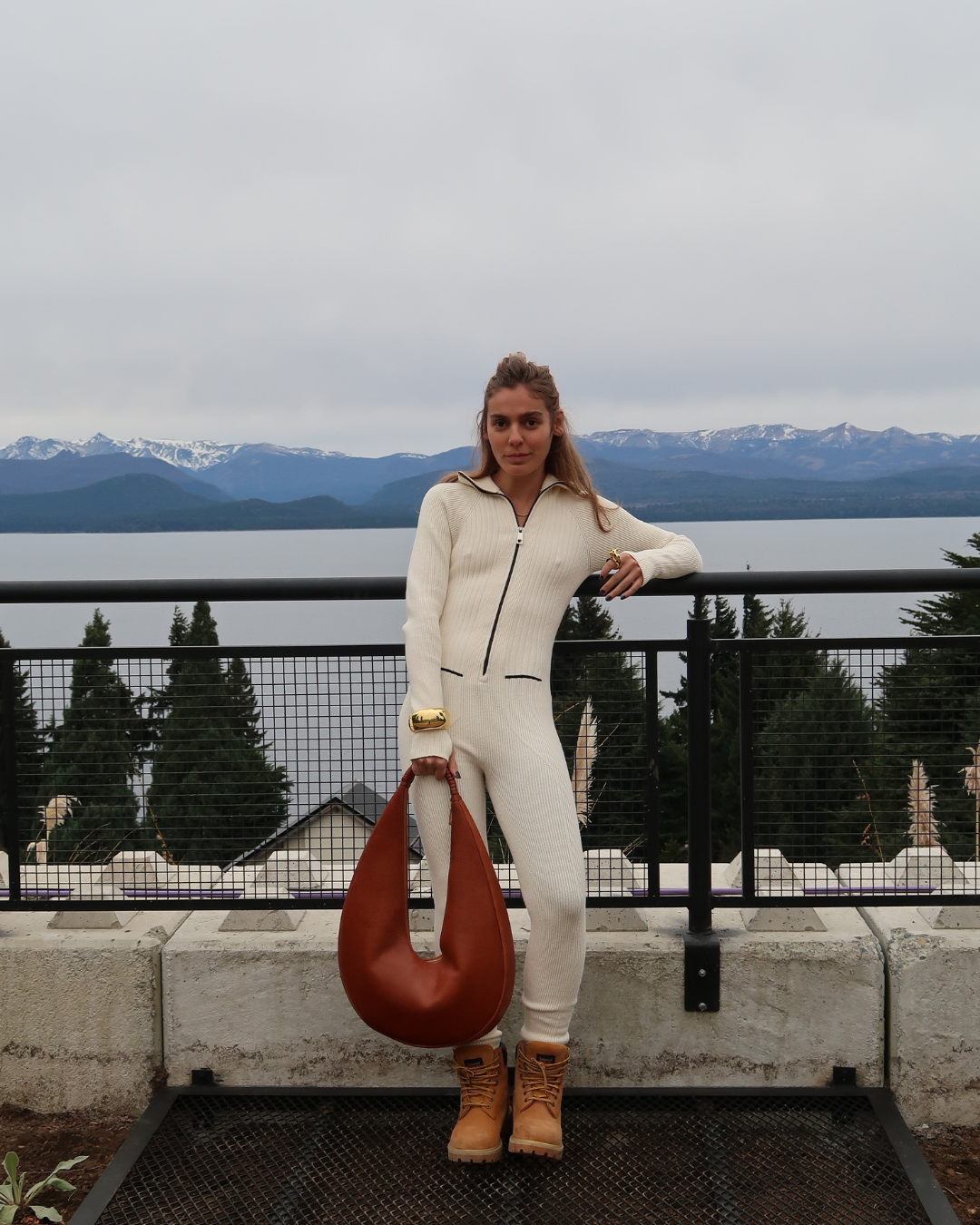 Pessoa usando macacão branco e botas, segura bolsa marrom em cenário de montanhas; tendências de alto inverno.