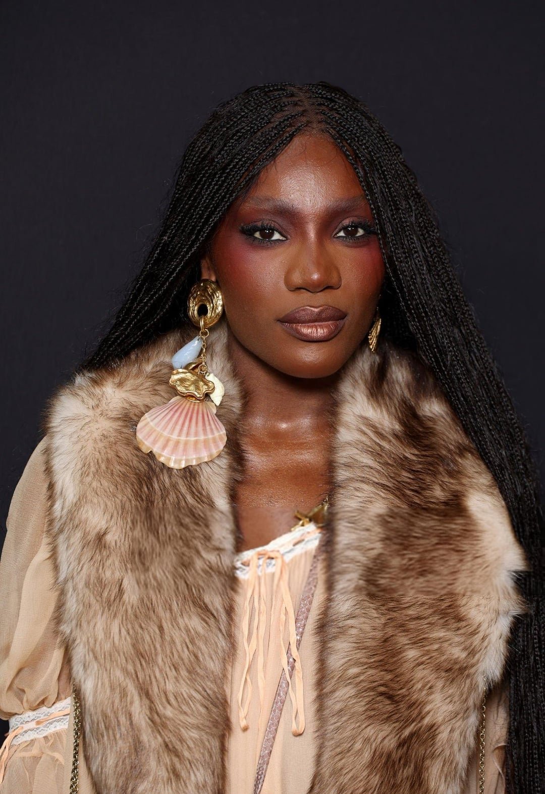 Usa brinco grande de concha em estilo acessório boho, vestida com colete de pele e blusa bege.