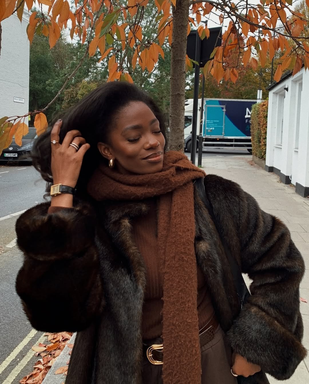 Mulher sorrindo em uma rua arborizada, vestindo casaco de pele e cachecol marrom, usando acessórios quentinhos.