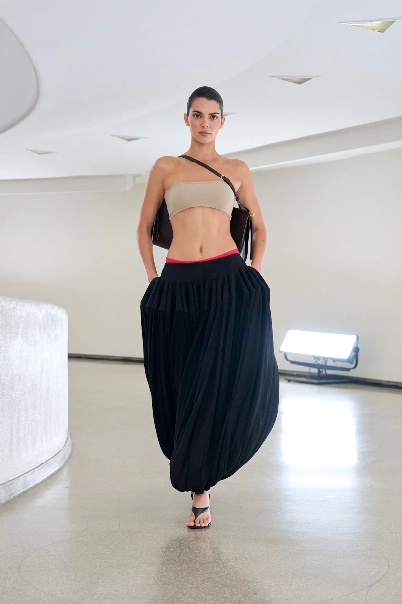 Modelo veste calça balonê preta com top bege, desfilando em ambiente moderno e minimalista.