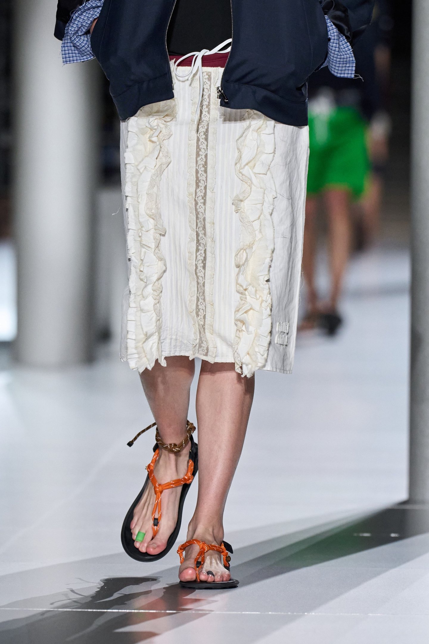 Sandália laranja com enfeite no tornozelo e anel no dedo do pé, combinada com saia branca rendada.