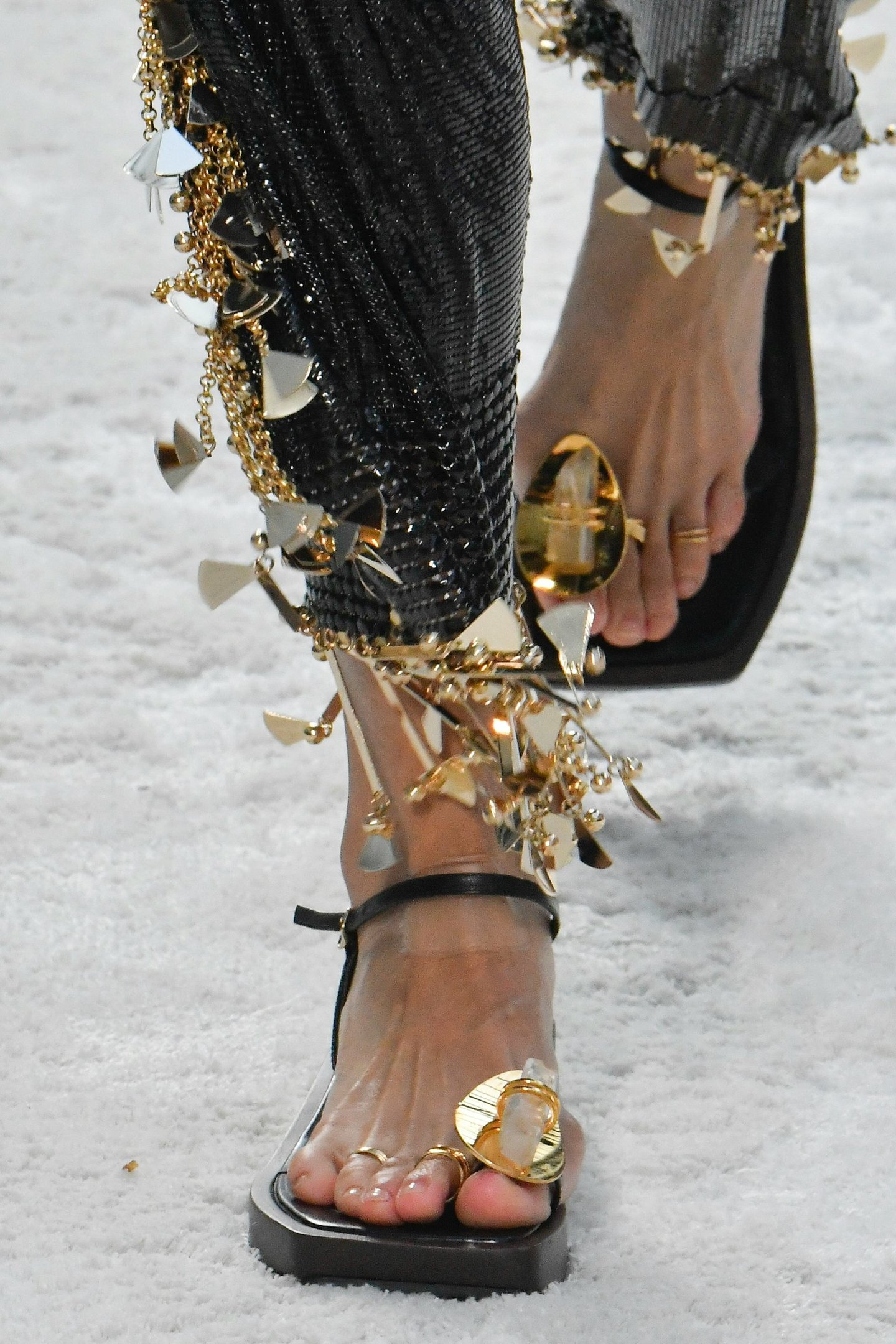 Sandália preta com ornamentos dourados, destacando anéis no dedo do pé e calça com franjas metálicas douradas.