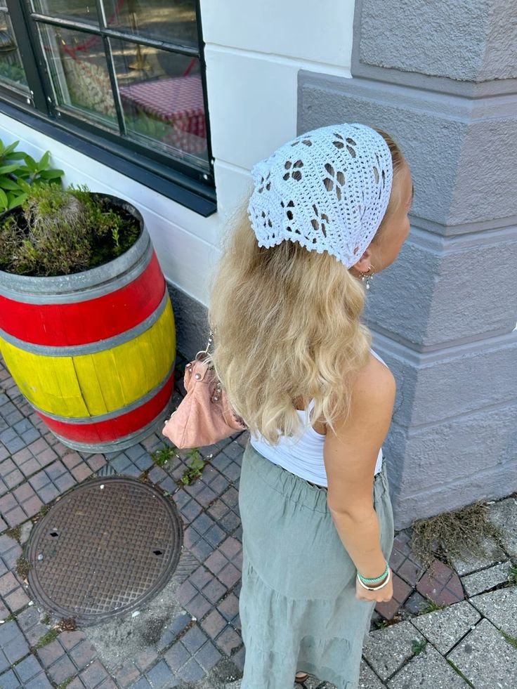 Mulher de cabelo loiro usa uma bandana de crochê branca, em pé próxima a um barril colorido.