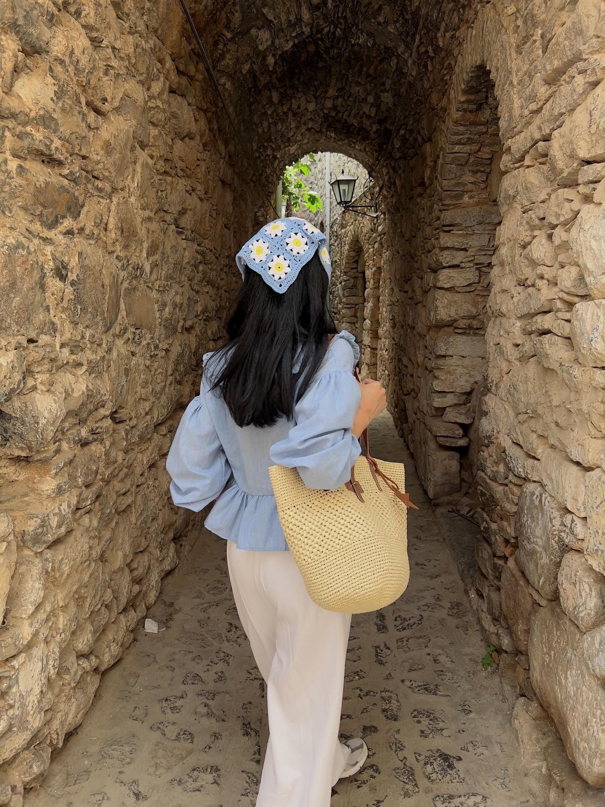 Pessoa em túnel de pedra usando blusa azul, bandana de crochê com flores e bolsa de palha.