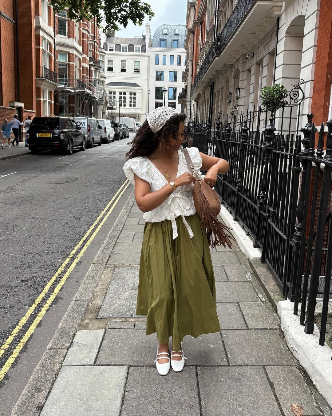 Mulher com bandana de crochê em rua arborizada, usando blusa branca e saia verde. Verificando bolsa marrom com franjas.