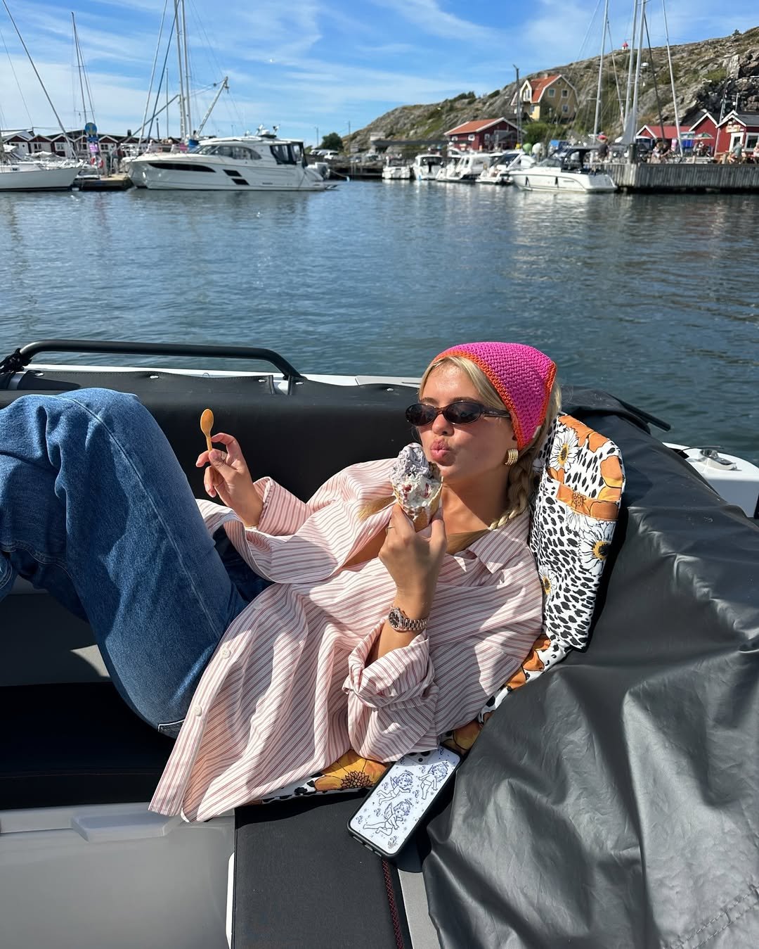Pessoa relaxando em um barco, com bandana de crochê rosa, óculos escuros e sorvete em mãos. Barcos ao fundo.