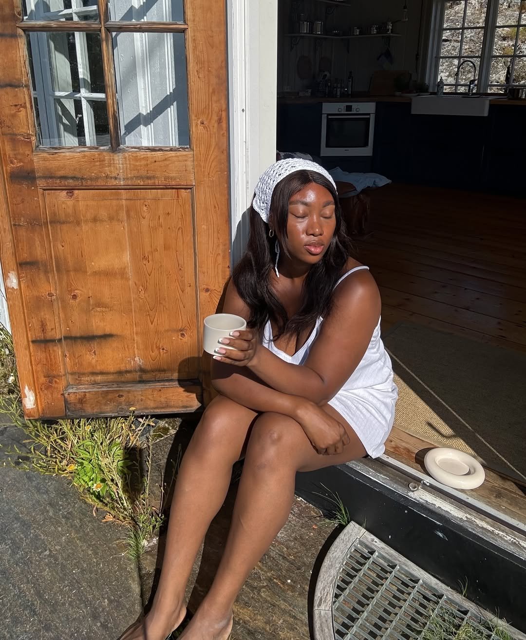 Mulher sentada ao sol, usando bandana de crochê branca e segurando uma xícara, em frente a uma porta de madeira.