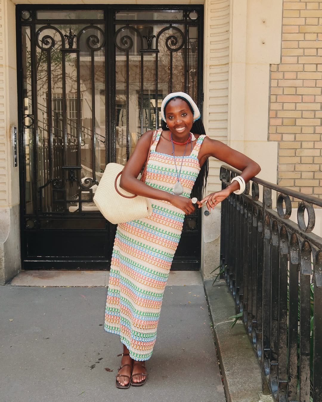 Pessoa usando vestido colorido longo e bandana de crochê branca, sorrindo em frente a uma porta de ferro.