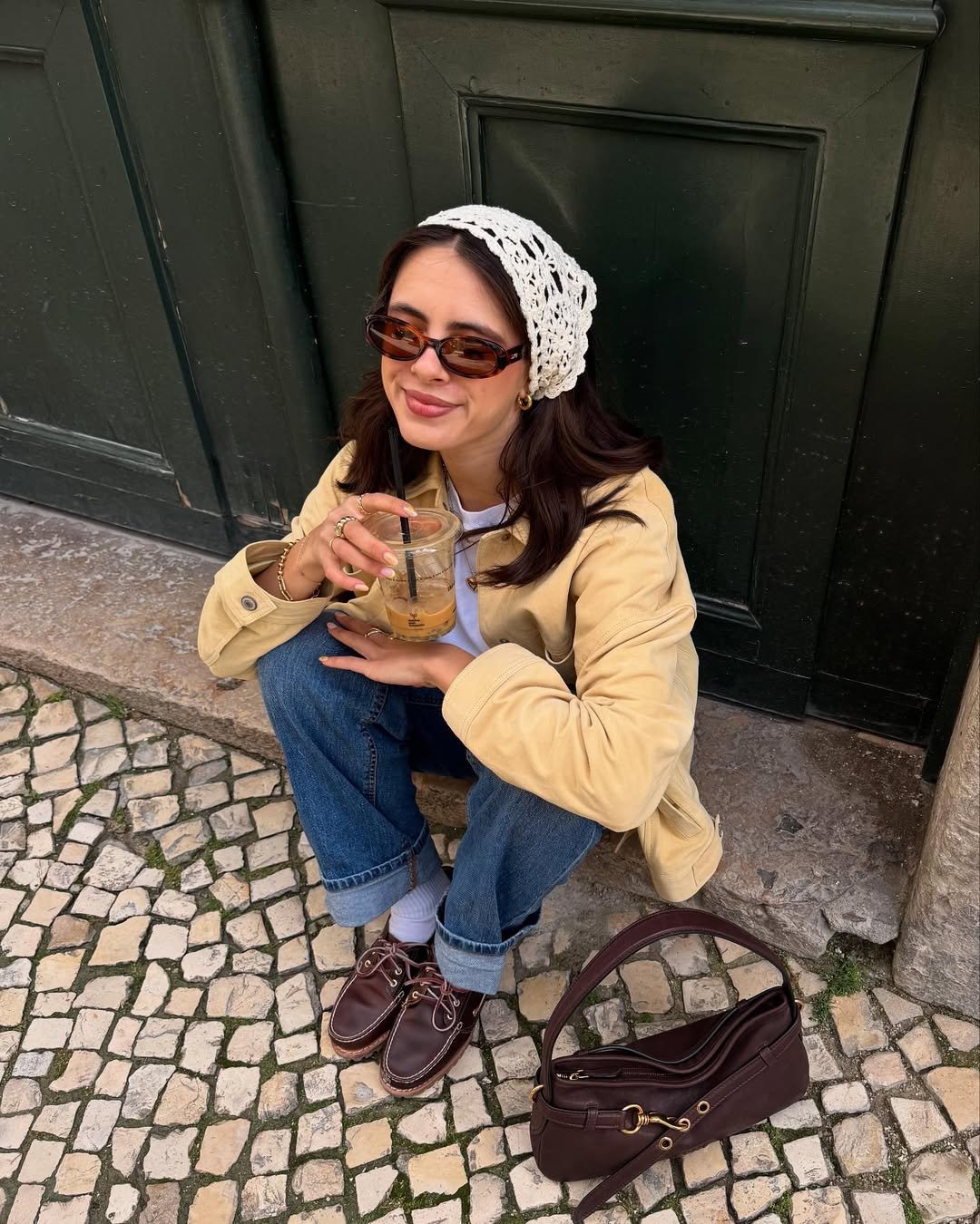 Mulher sorrindo, usando bandana de crochê branca e óculos escuros, segura bebida sentada em frente a uma porta verde.