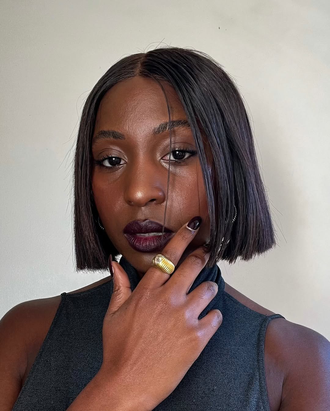Mulher com batom vinho e cabelo curto, usa anel dourado e toca o rosto.