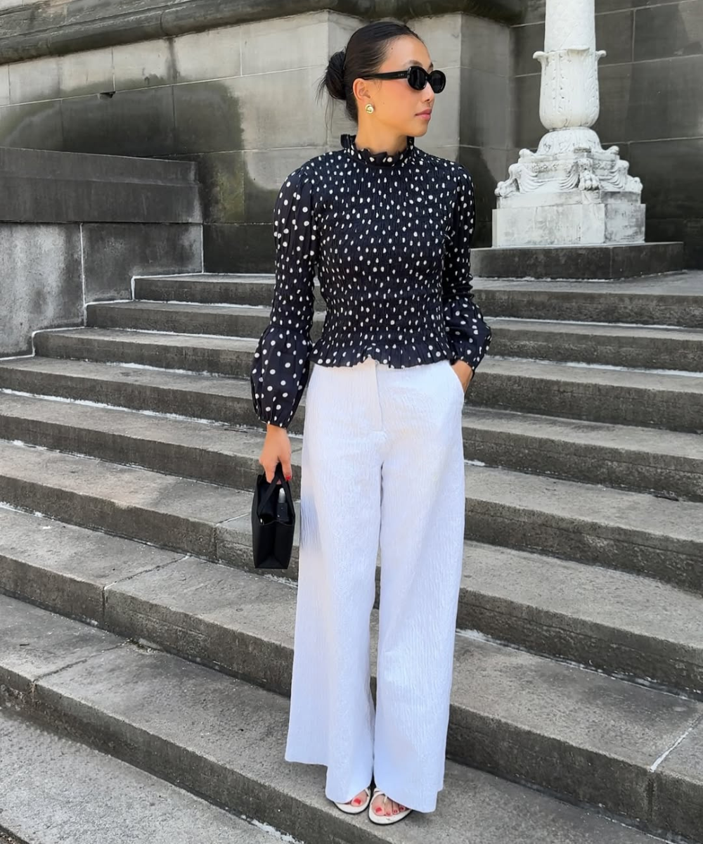 Pessoa de óculos escuros usando blusa de poá preta e calça branca em uma escadaria.