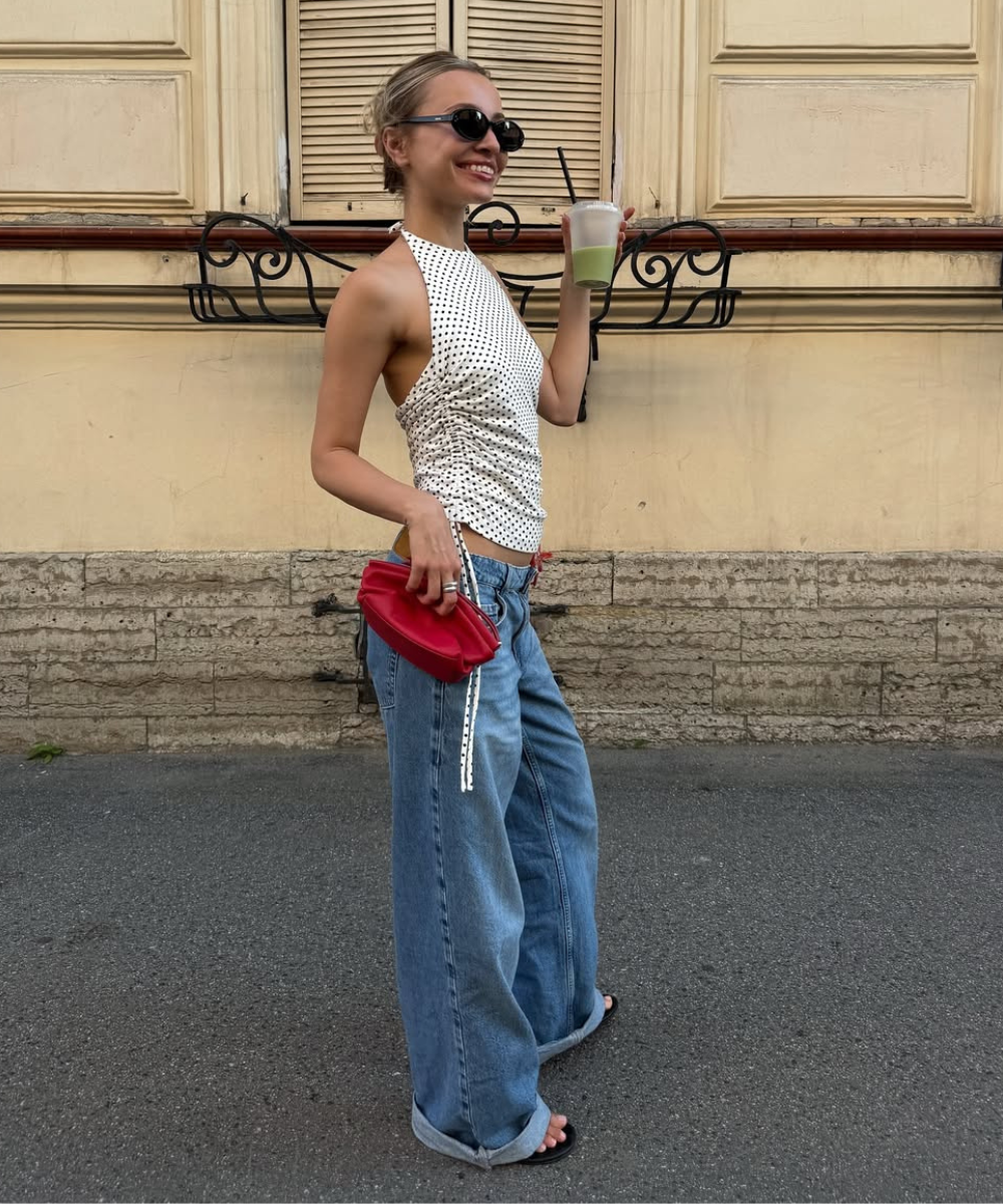 Mulher sorridente com blusa de poá e calça jeans larga segura bebida e bolsa vermelha em rua com parede colonial.