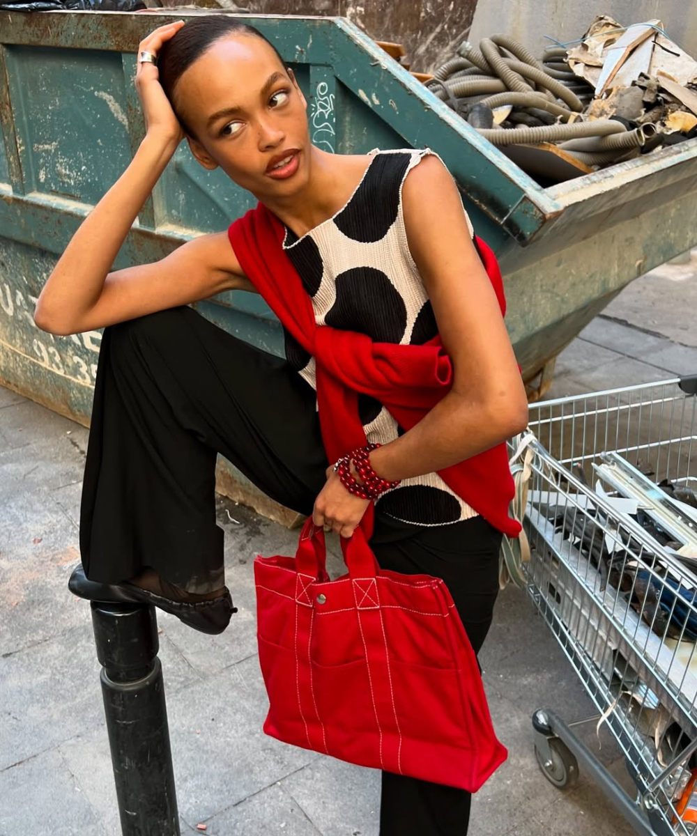 Pessoa usando blusa de poá preto e branco, segurando bolsa vermelha ao lado de um contêiner.