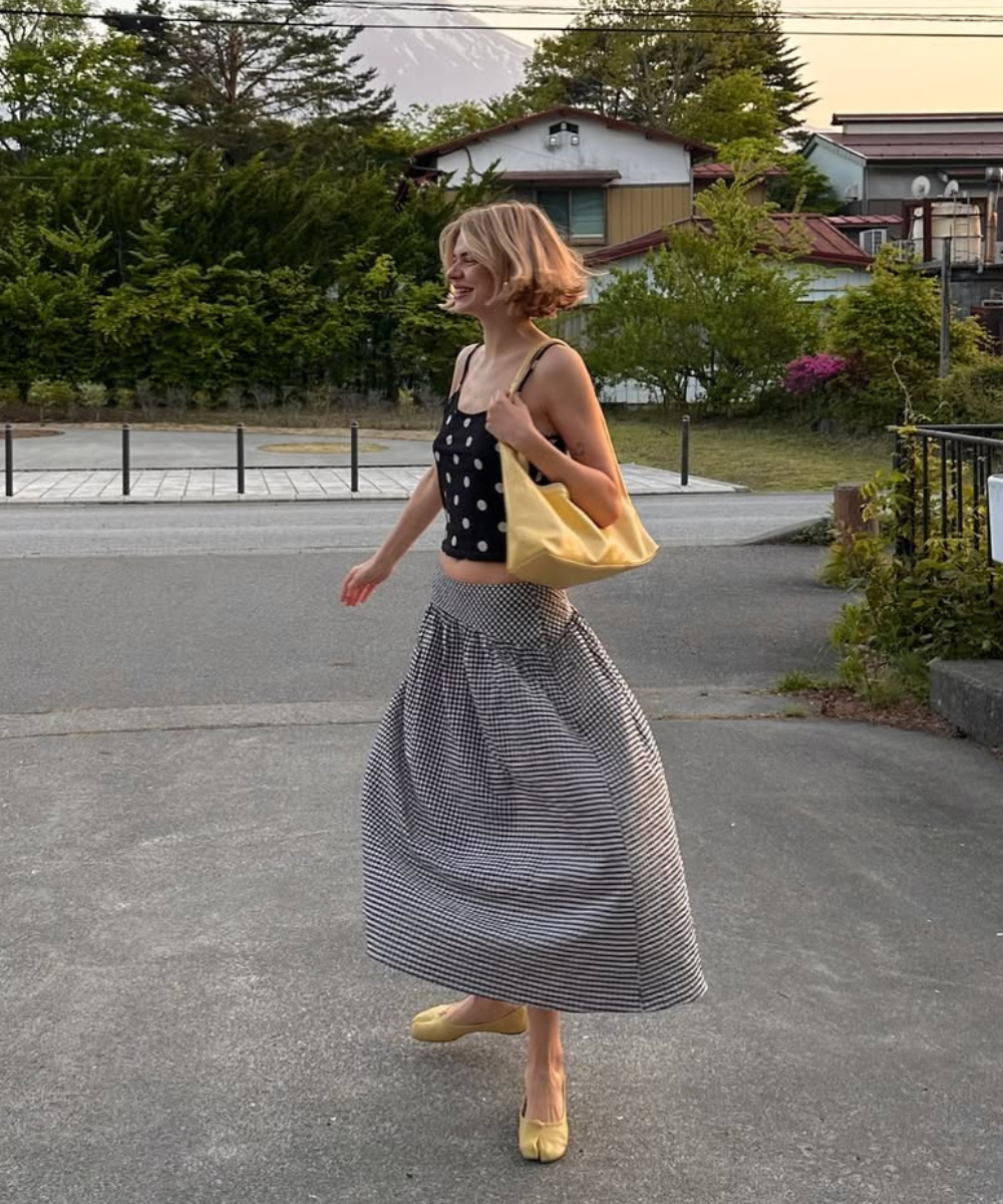 Mulher sorrindo, usando blusa de poá, saia listrada, bolsa e sapatos amarelos, andando ao ar livre.