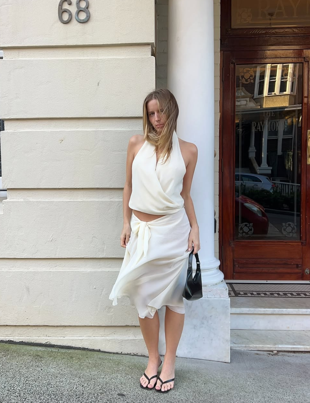 Pessoa com blusa frente única branca e saia amarrada, em frente a um prédio.