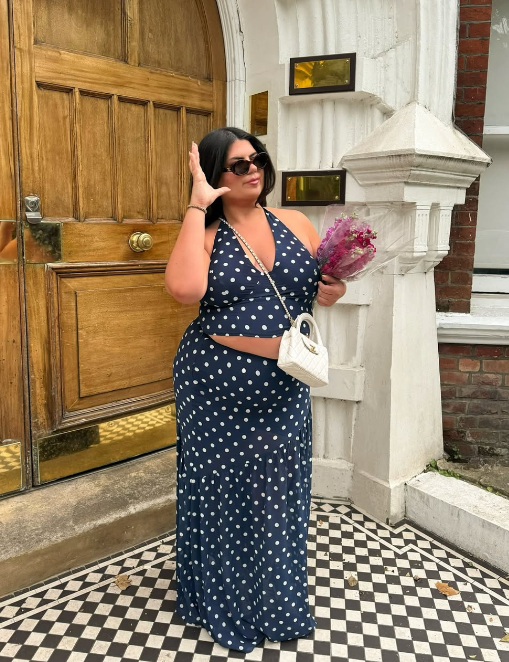 Mulher com vestido de bolinhas segurando flores, em frente a uma porta de madeira elegante.