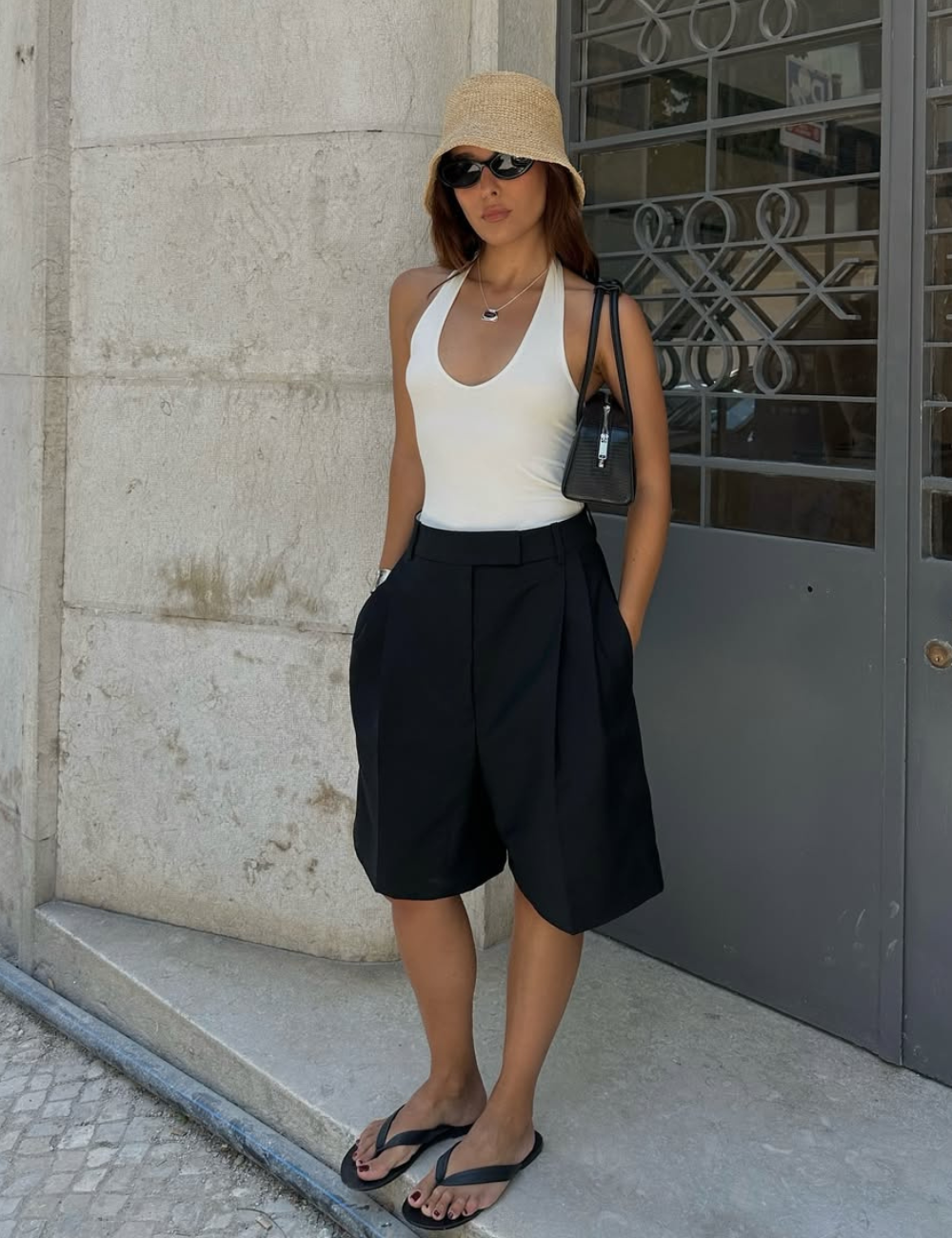 Pessoa com blusa frente única branca e chapéu bege, calça preta larga e chinelo, em frente a uma porta cinza.