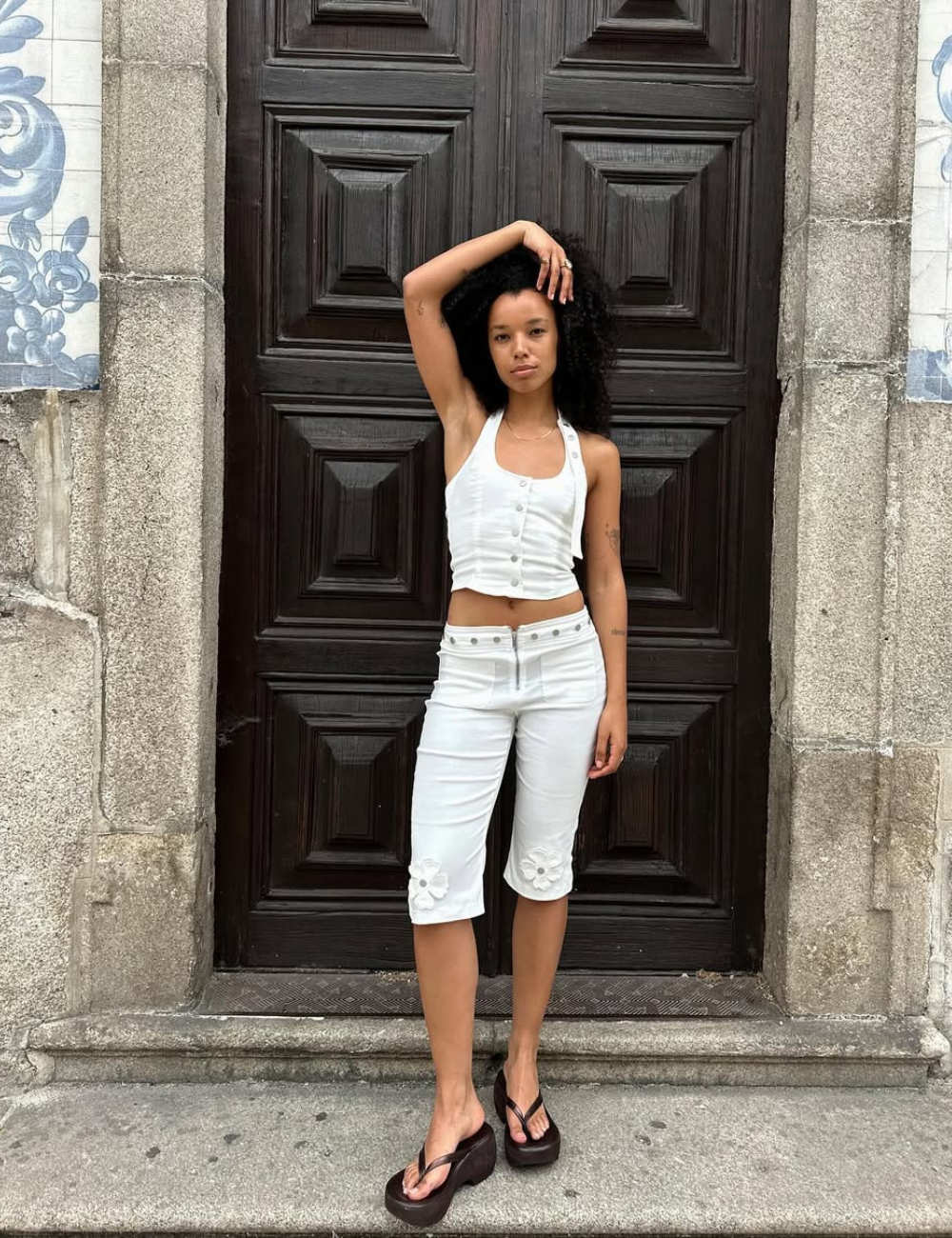 Pessoa usando blusa frente única branca, posando em frente a uma porta de madeira elaborada, usando bermuda branca e sandálias plataformas.