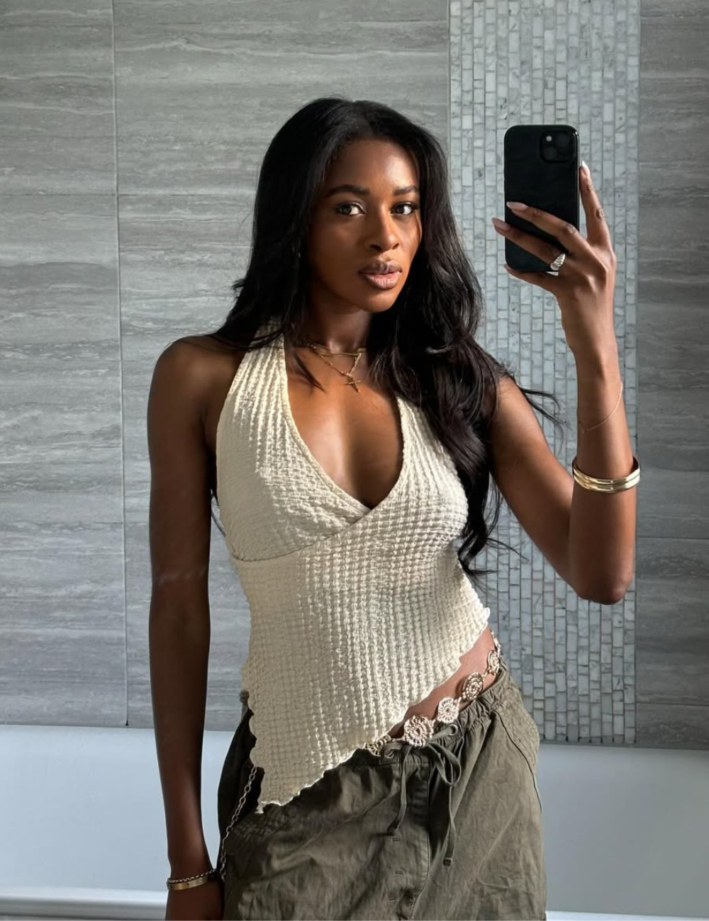 Pessoa usando blusa frente única bege, posando em um ambiente interno com parede de azulejos ao fundo.