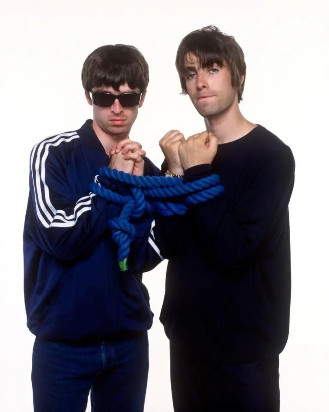 Dois homens com trajes casuais unidos por corda em estilo associável ao britpop.