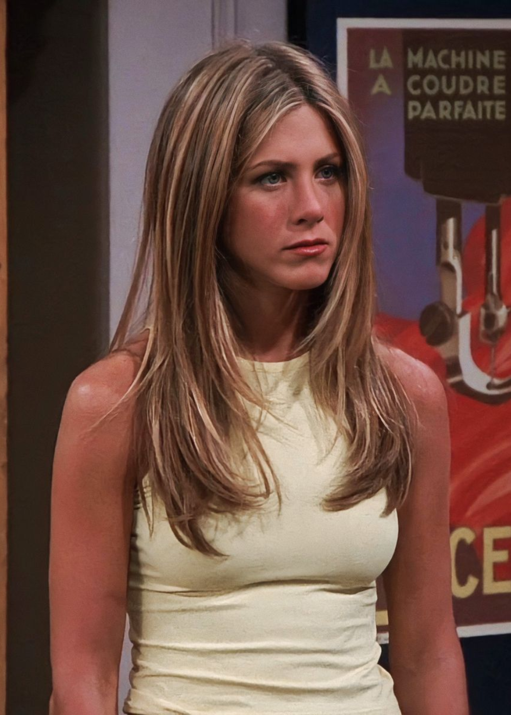 Pessoa com cabelo médio ondulado semelhante ao estilo da Rachel Green, vestindo blusa clara. Ao fundo, pôster colorido.