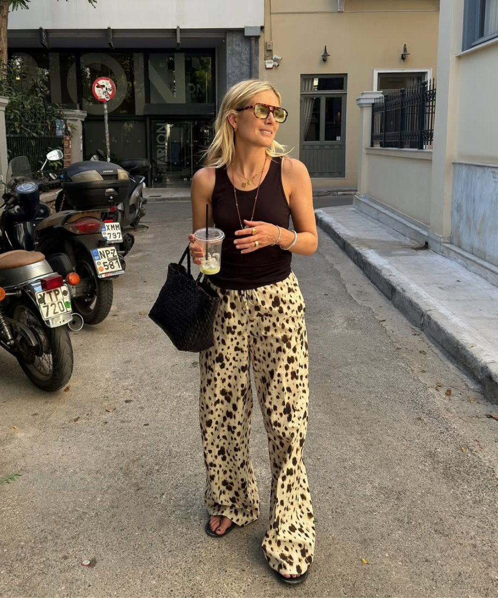 Pessoa usando calça estampada com chinelo em rua urbana. Ela segura bebida e bolsa preta.