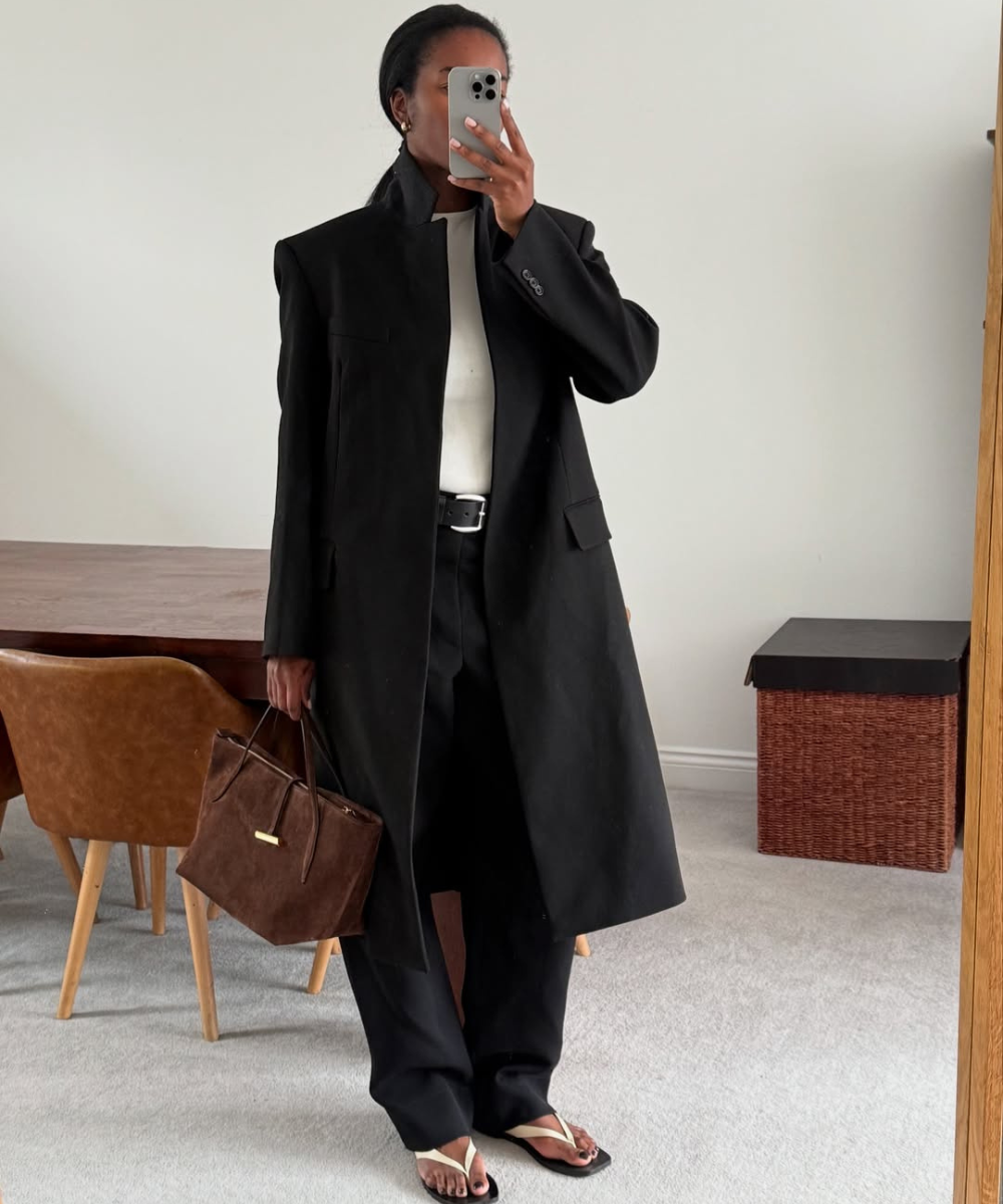 Mulher de casaco preto, calça com chinelo claro e bolsa marrom, segurando celular em sala com móveis de madeira.