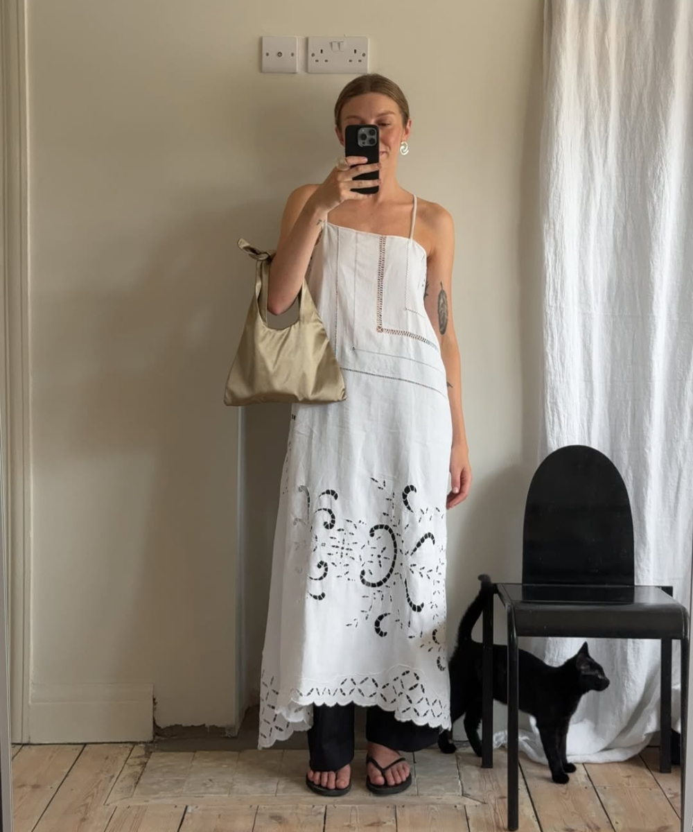 Pessoa usando vestido branco sobre calça com chinelo, segurando uma bolsa. Ao lado, um gato preto e uma cadeira.