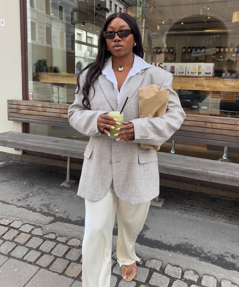 Pessoa vestindo blazer cinza e calça com chinelo, segurando bebida verde em frente a café na rua.