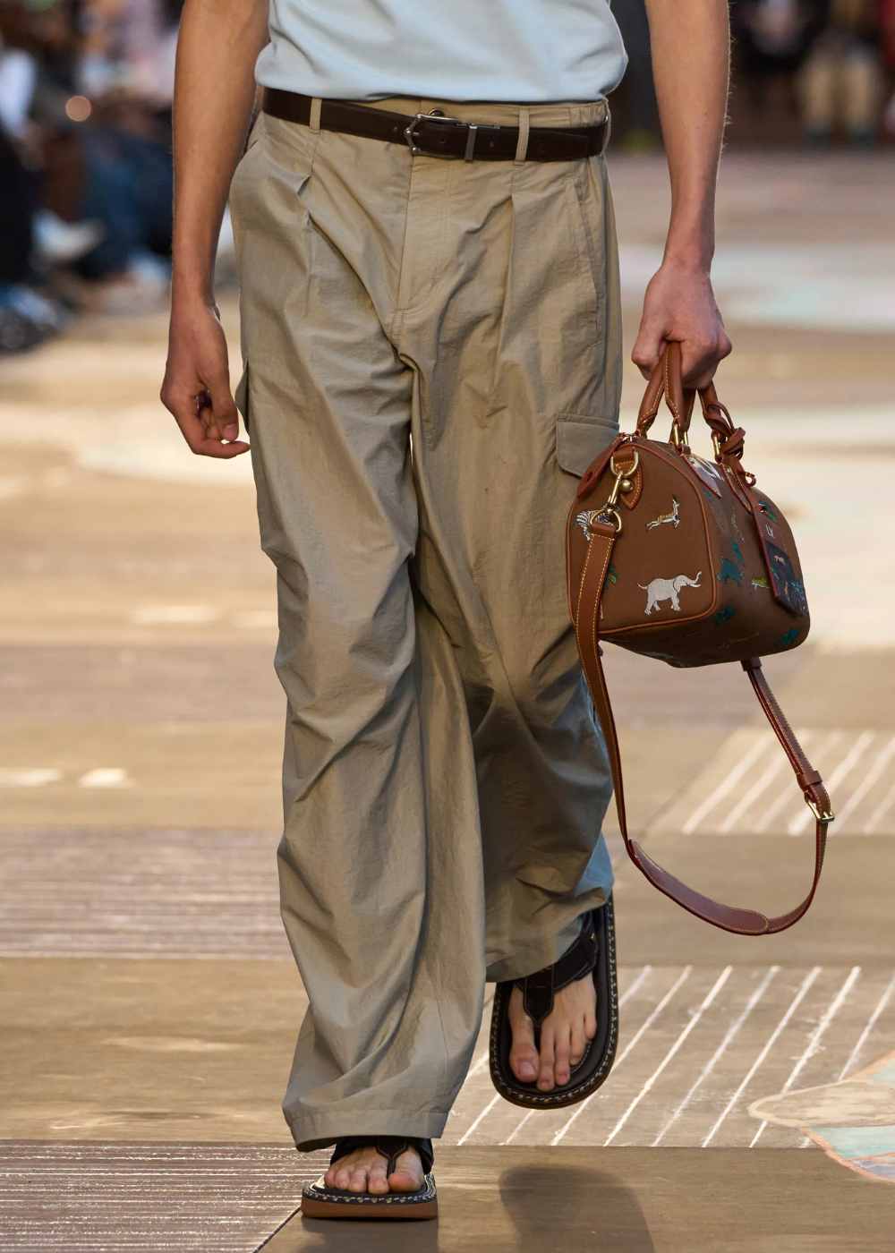 Calça bege com chinelo preto e bolsa marrom com estampas.