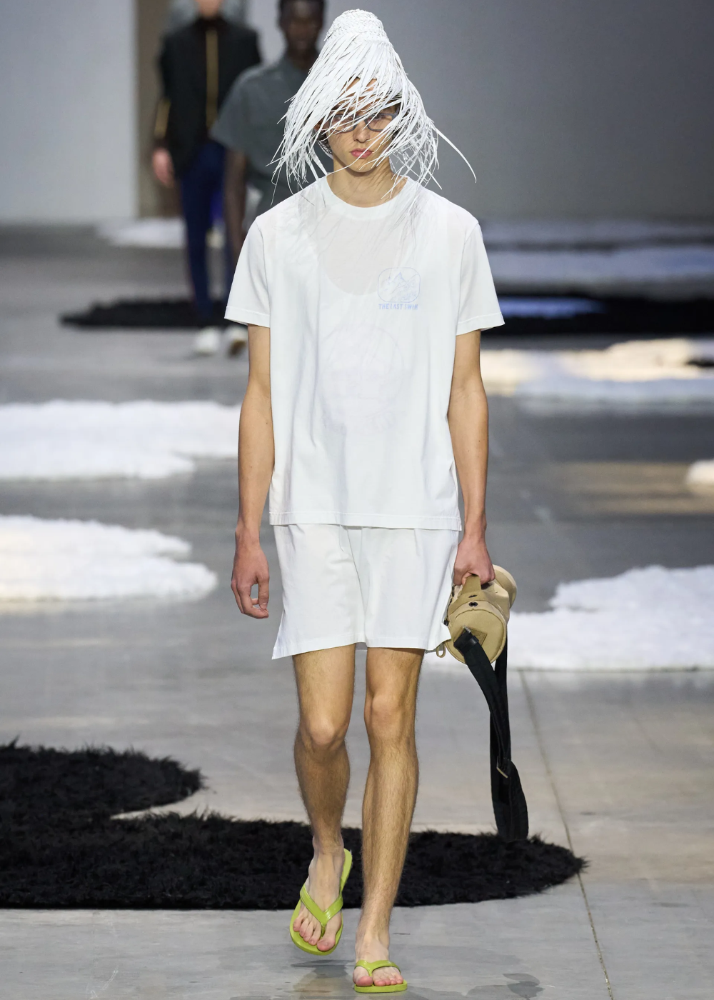 Modelo veste branco com chapéu estilizado e segura calça, usando chinelo verde.