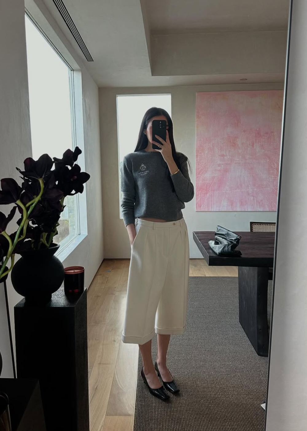 Pessoa com suéter cinza e calça culotte branca, espelho ao fundo, flores escuras à esquerda.