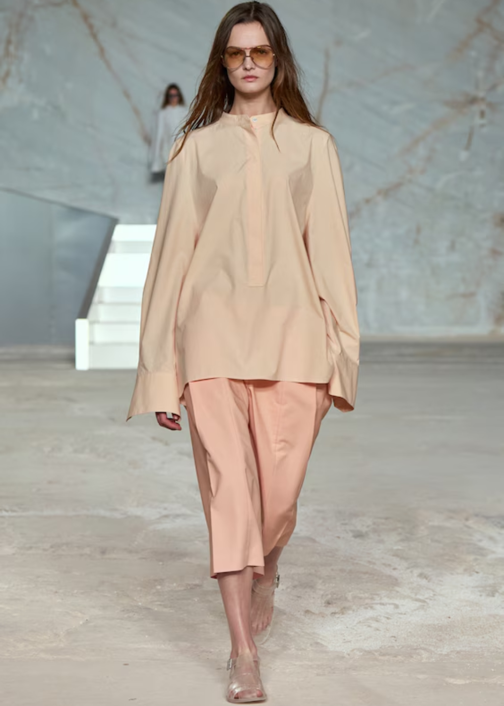 Modelo desfila com calça culotte e blusa de tom nude em passarela minimalista, usando óculos escuros.