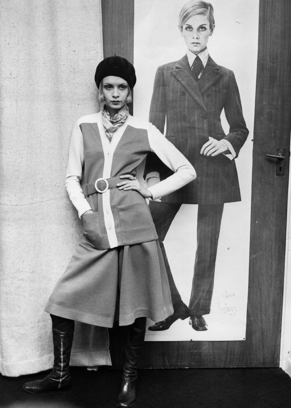 Modelo usa calça culotte com blazer e botas, em frente a um pôster vintage.