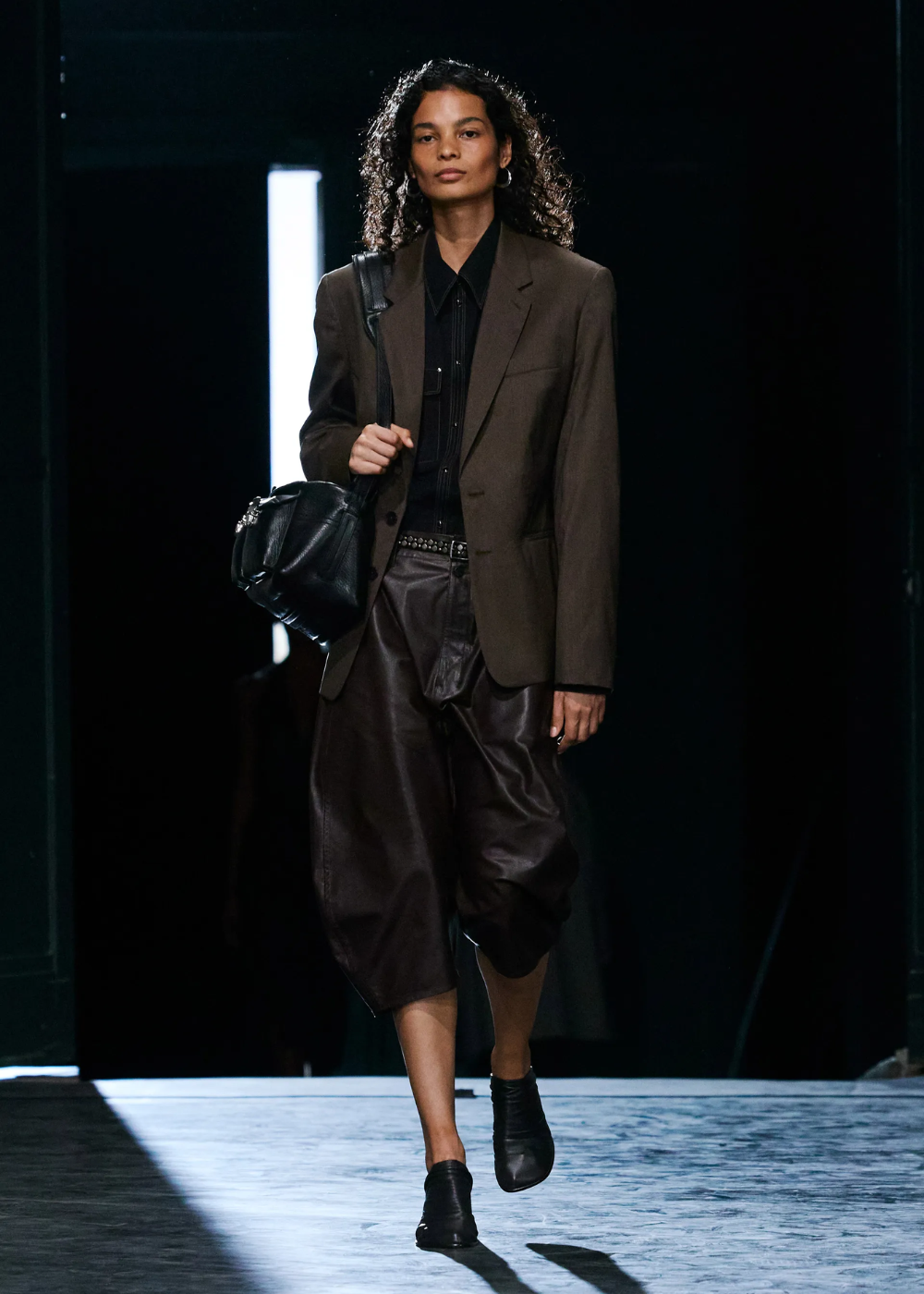Modelo desfilando com blazer marrom e calça culotte larga, segurando uma bolsa preta.