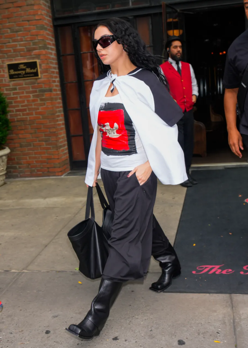 Pessoa veste calça culotte preta, camiseta estampada vermelha e capa branca, carregando bolsa preta.