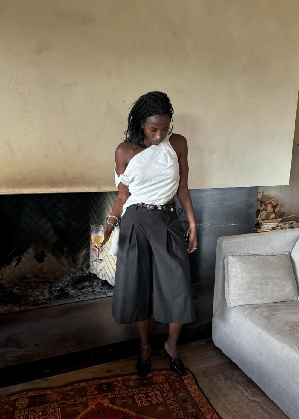 Pessoa usando calça culotte preta e blusa branca, segura taça em ambiente interno acolhedor.
