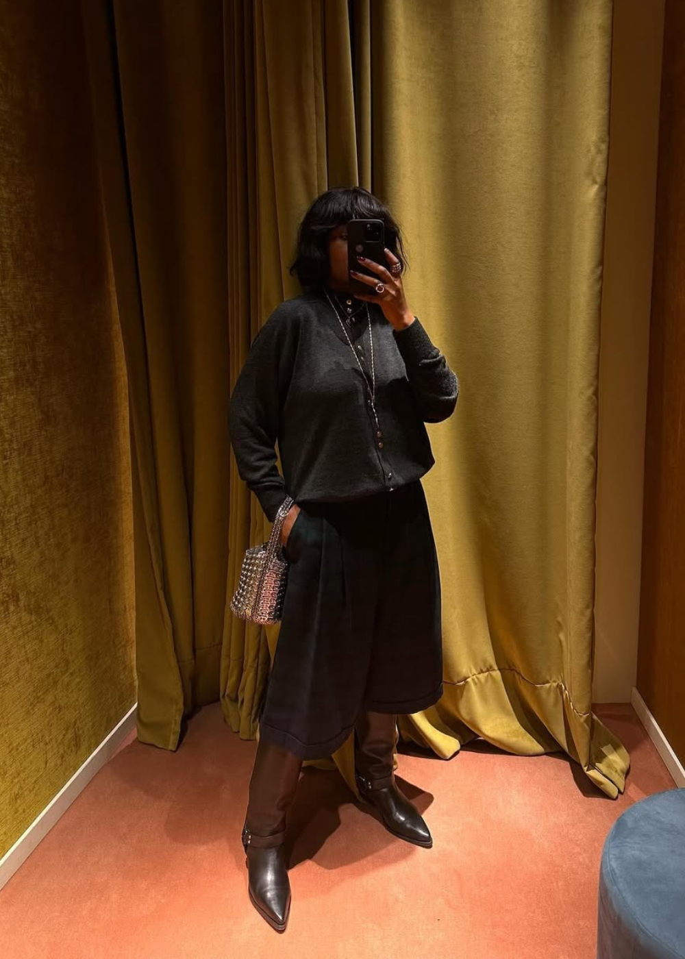 Pessoa com blusa preta e calça culotte escura posa em frente ao espelho com cortina dourada ao fundo.