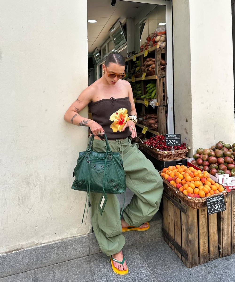 Loja de frutas com mulher usando top colorido, calça de nylon verde e chinelos amarelos, segurando bolsa verde.