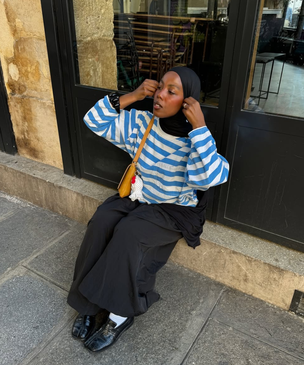 Pessoa sentada na calçada, ajustando hijab. Usa camiseta listrada azul e calça preta. Bolsa amarela pendurada.