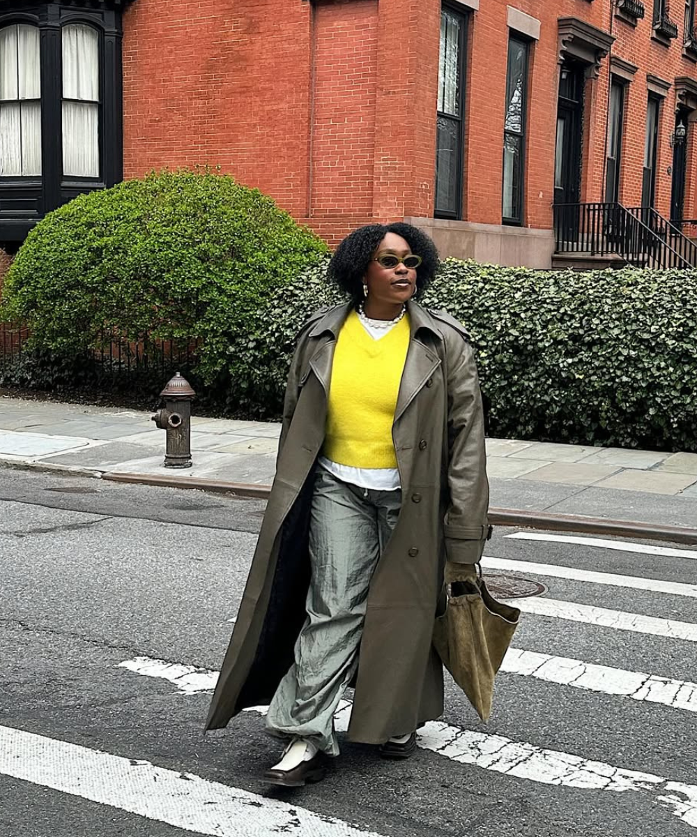 Pessoa atravessa rua com calça de nylon cinza, suéter amarelo e casaco longo, carregando bolsa marrom.