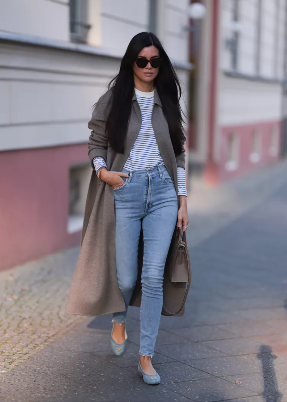 Mulher usando casaco longo, blusa listrada e calças jeans tendência. Segura bolsa cinza e veste sapatilhas.