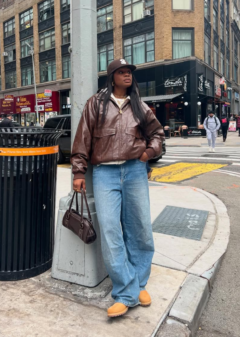 Pessoa com jaqueta de couro e calças jeans tendência em esquina de rua movimentada.