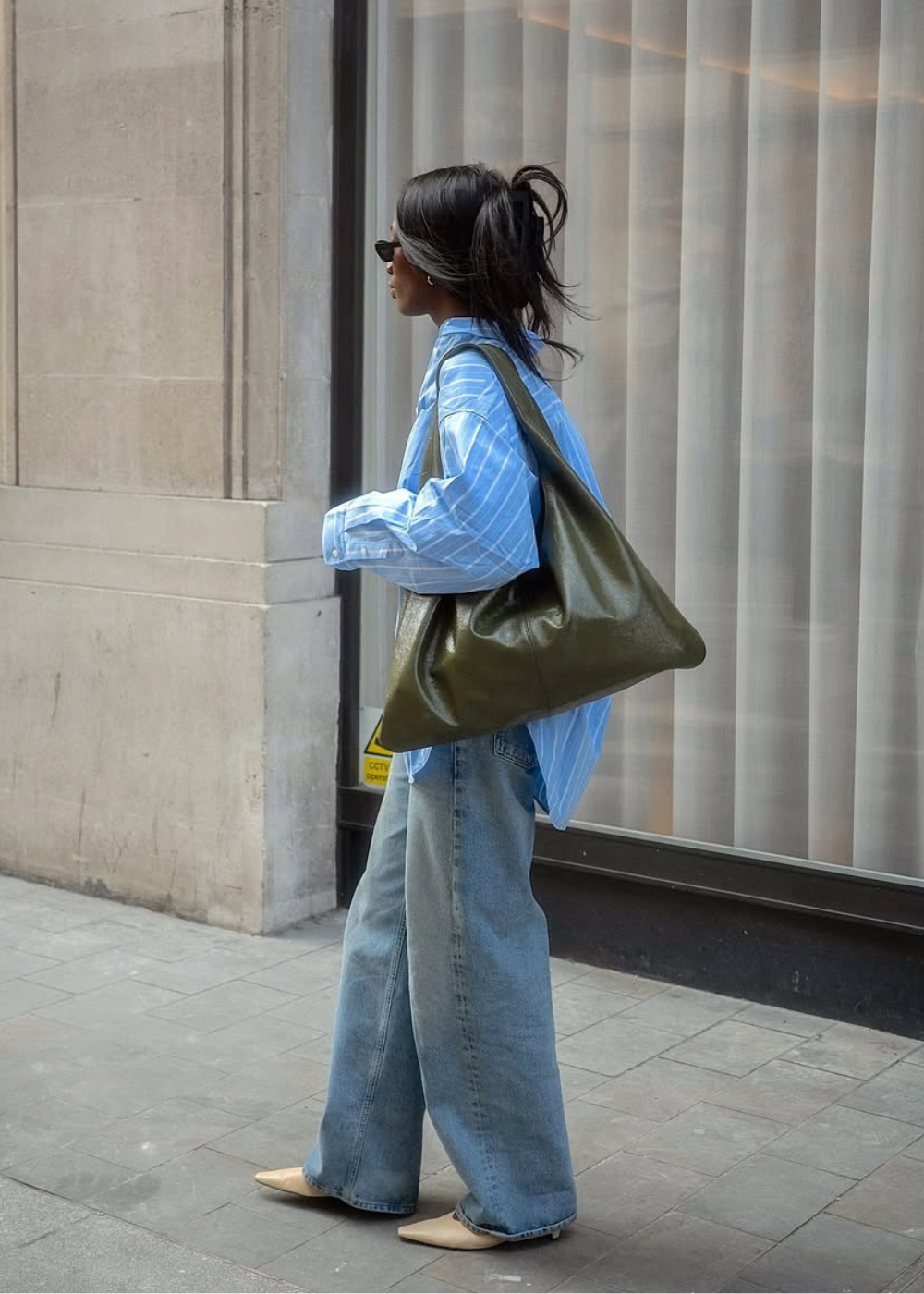Pessoa usando calças jeans largas, camisa azul e bolsa grande verde, caminhando na calçada.